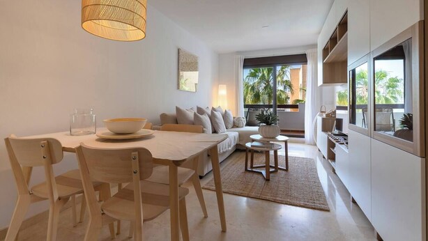 Een moderne woonkamer met een eethoek, een bank, een salontafel en uitzicht op palmbomen door grote ramen.