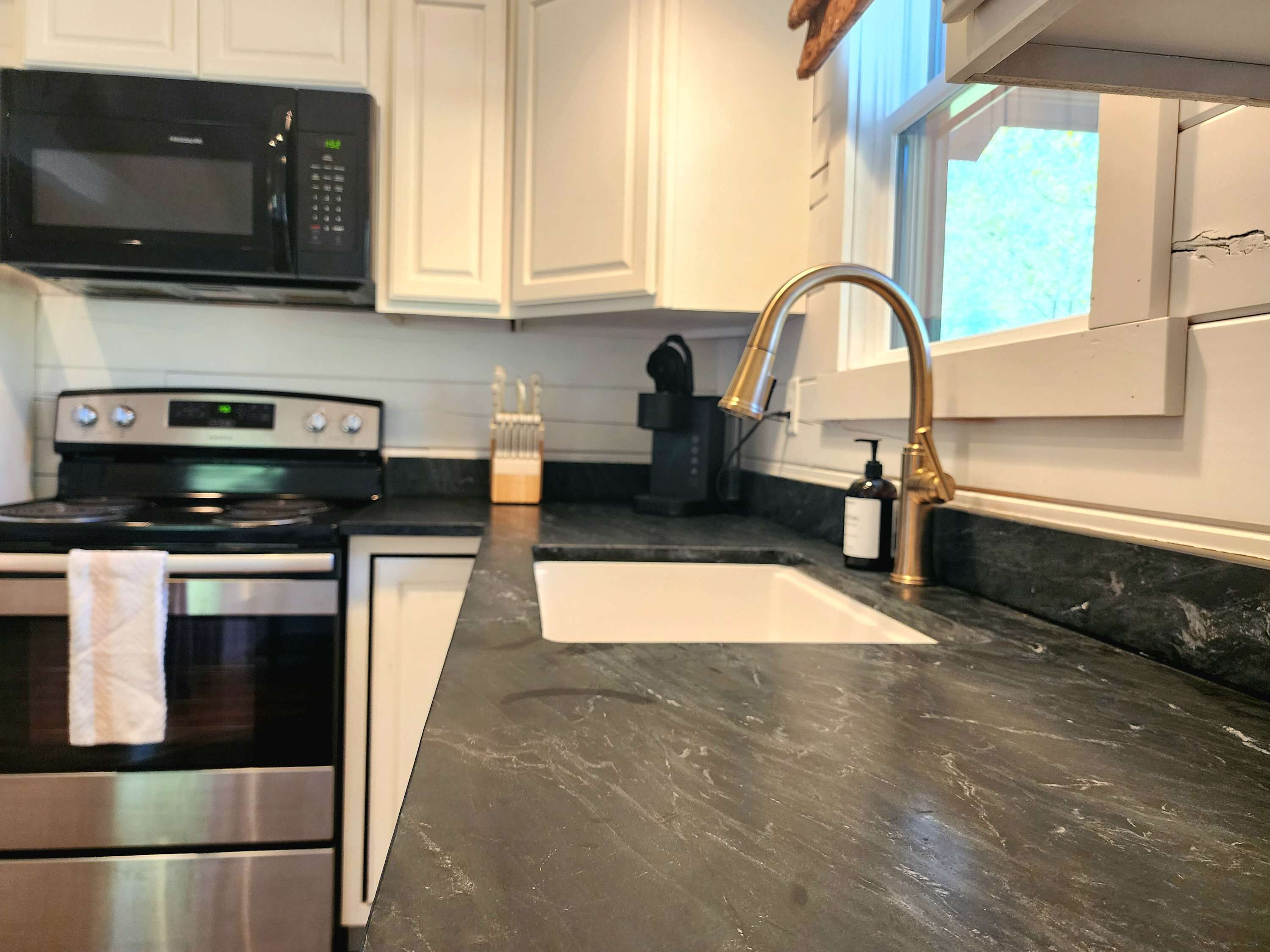 A kitchen with white cabinets, a black countertop, a stainless steel oven, a microwave, and a gold faucet.