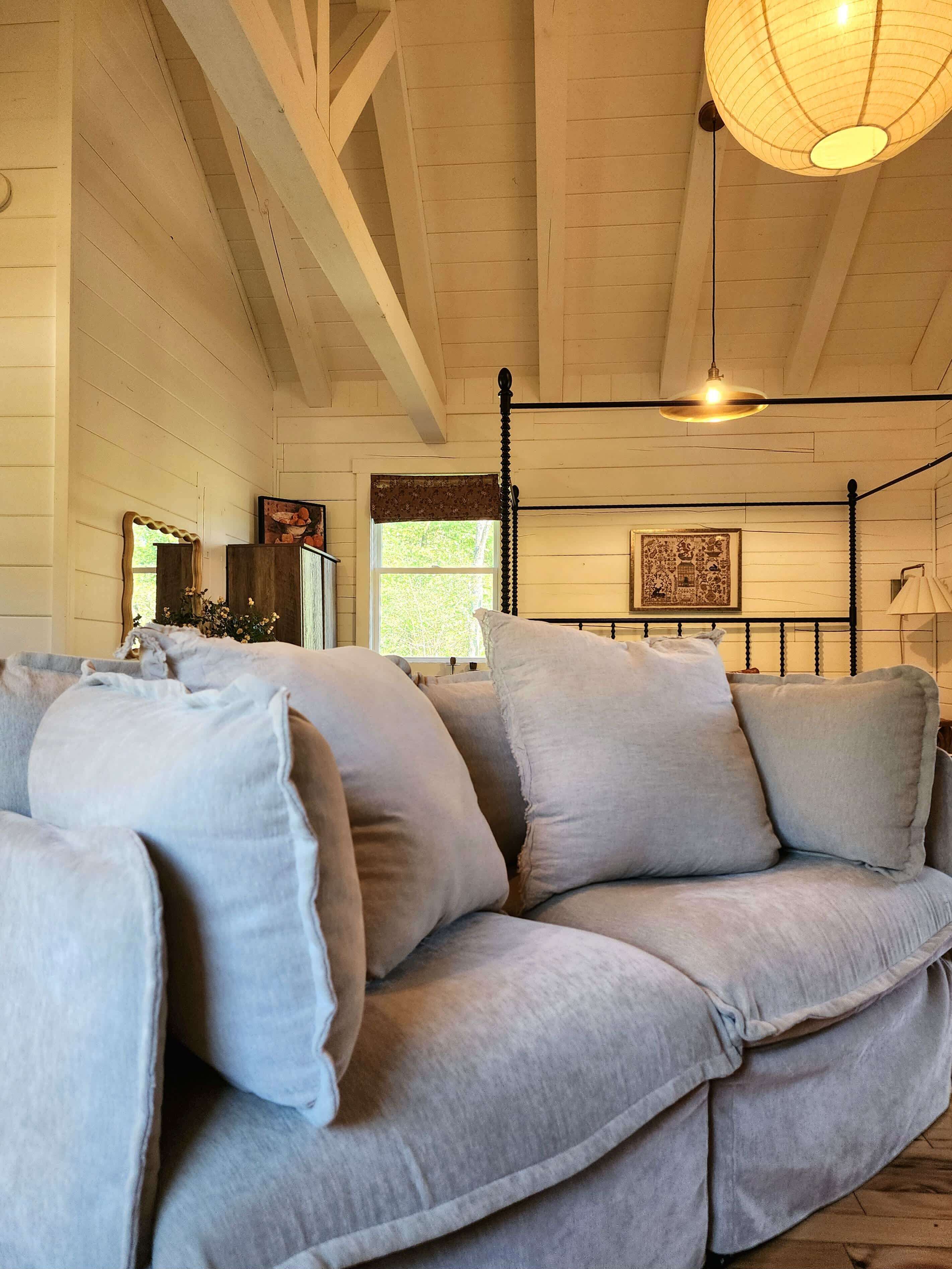 A room with a large grey sectional sofa, a wooden ceiling with exposed beams, a hanging light fixture, and a window with a view of greenery.