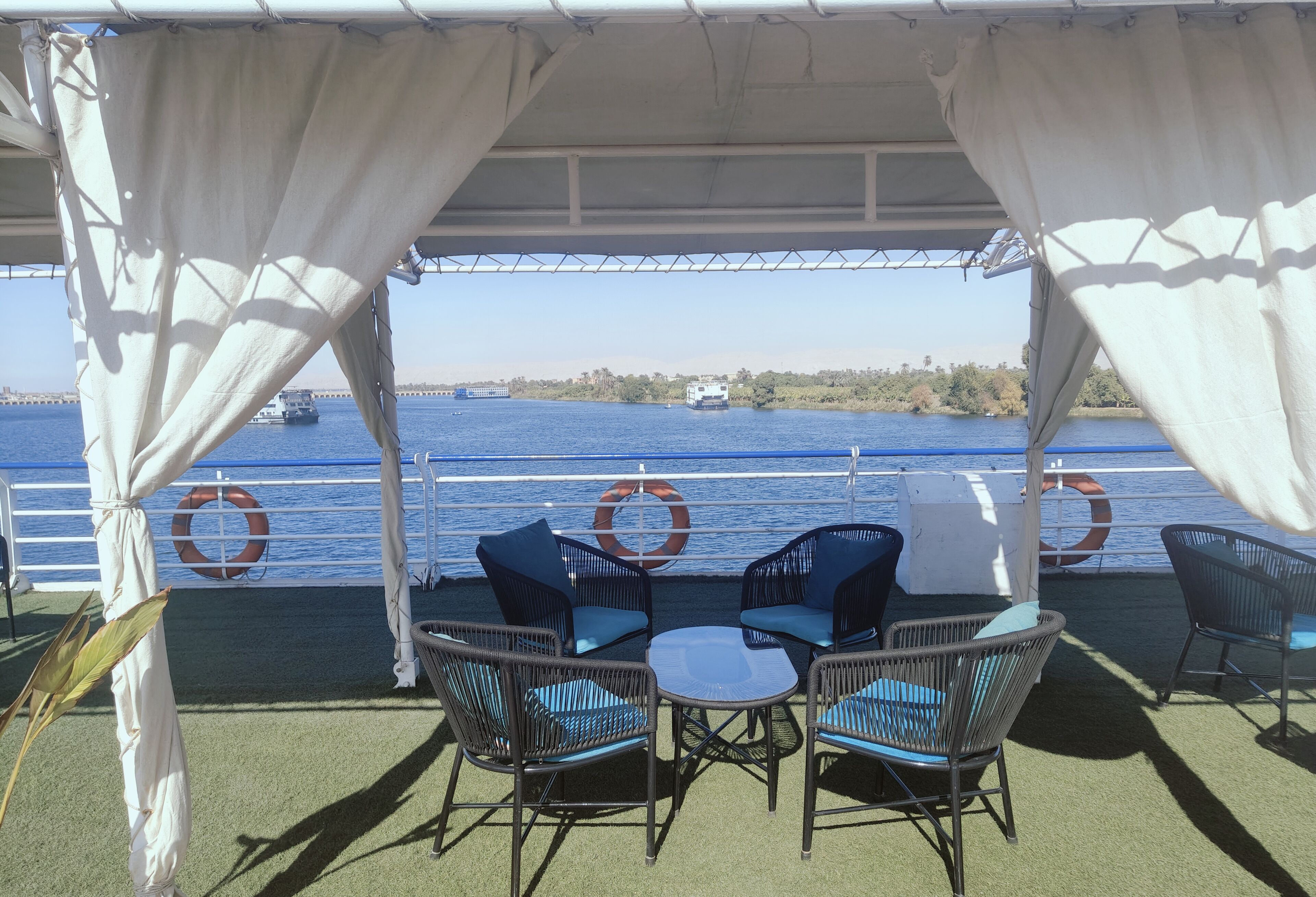 A covered outdoor seating area with wicker chairs and a small table, overlooking a body of water with boats and a distant shoreline.