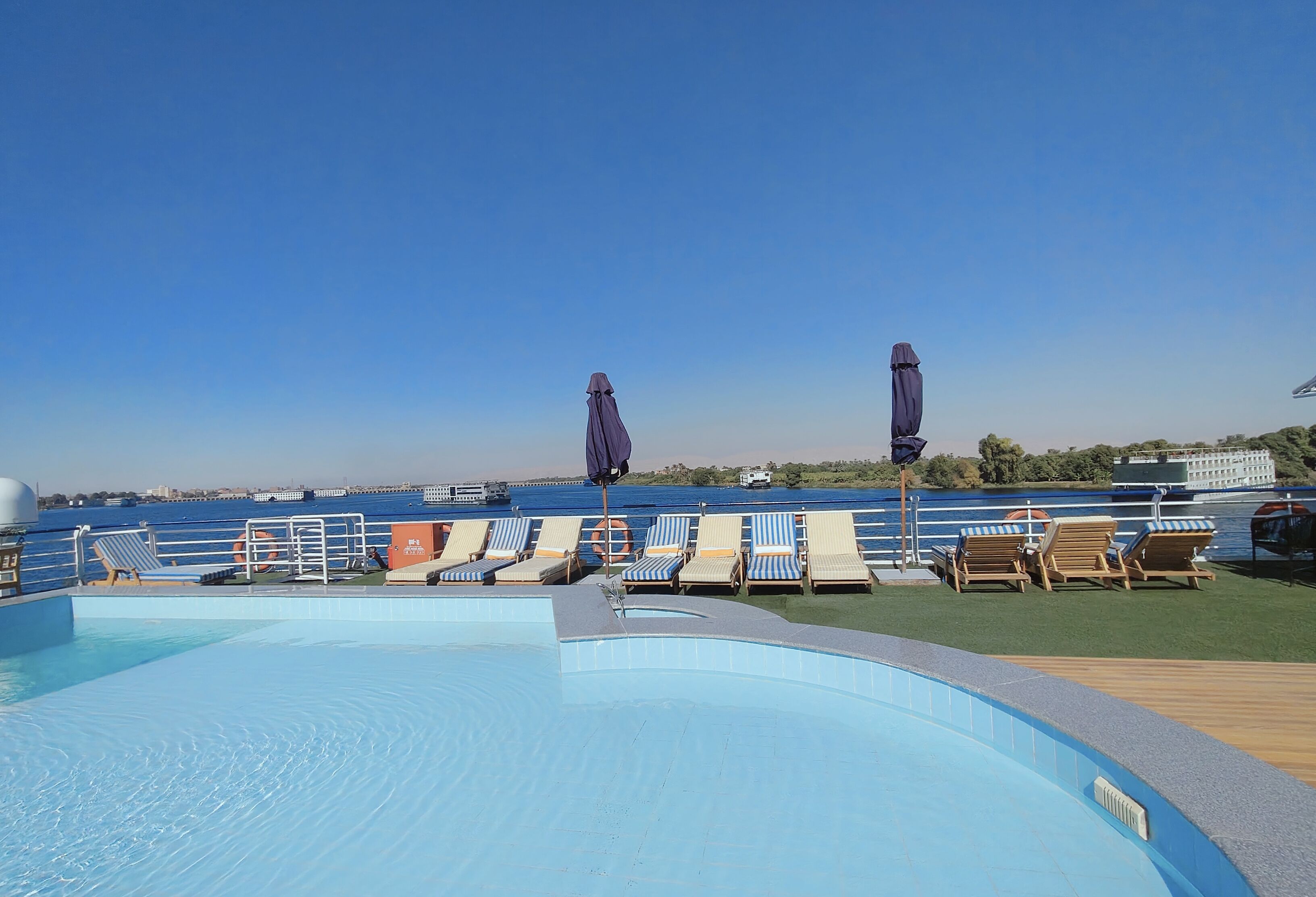 Zona de piscina con sillones de descanso y sombrillas, con vista a una masa de agua y una costa lejana.