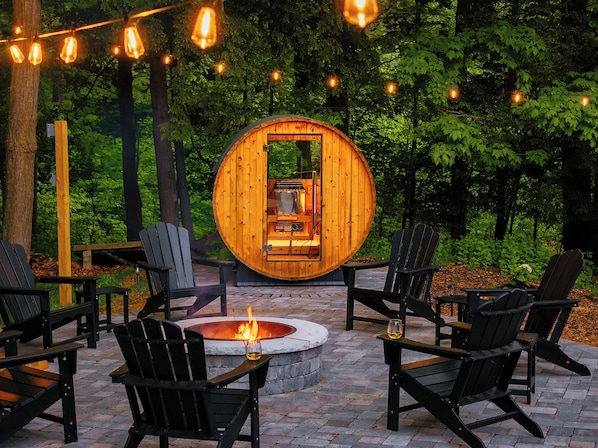 Una estructura circular de madera con un brasero en el centro, rodeada de sillas Adirondack negras y un patio de ladrillo.