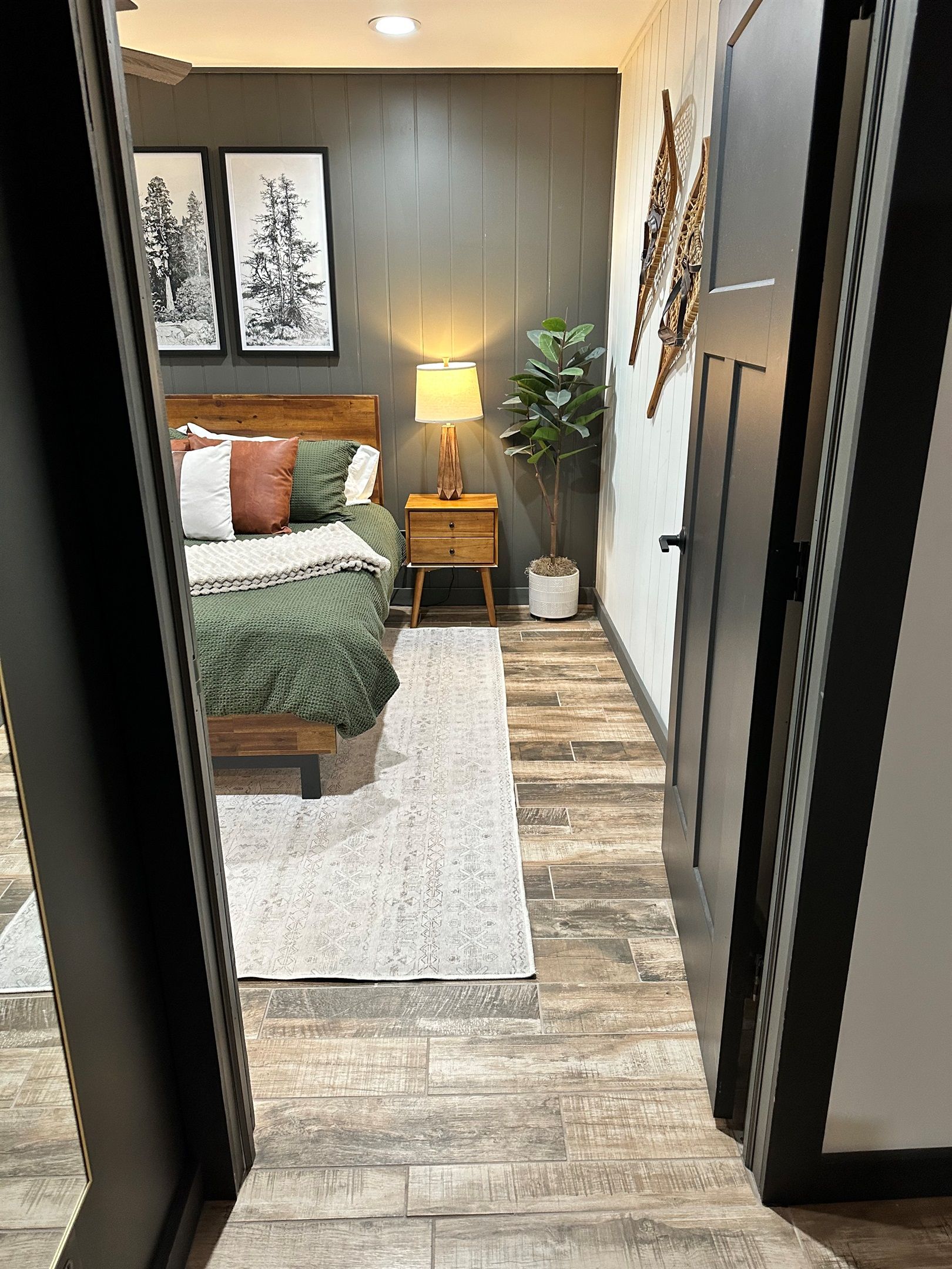 A bedroom with a bed, bedside table, lamp, potted plant, and framed pictures on the wall.
