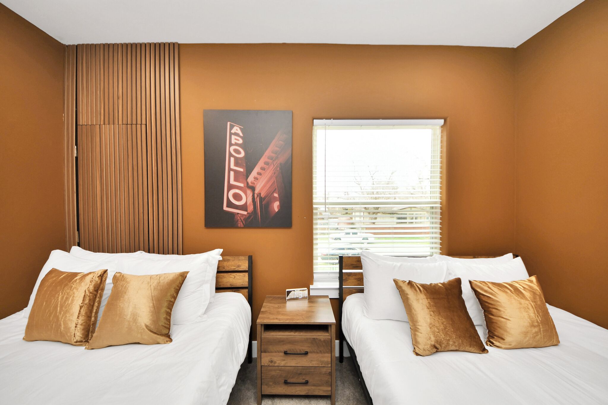 Een hotelkamer met twee bedden, een raam met zonwering, een houten nachtkastje en een lijst met een afbeelding aan de muur.