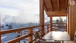 A wooden deck with a table and chairs, overlooking a mountainous landscape with buildings and a cloudy sky.