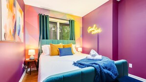 Une chambre d’hôtel moderne avec un lit de couleur turquoise, des œuvres d’art abstraites aux couleurs vives, un néon affichant le message « Bonne ambiance » et une fenêtre avec des rideaux.
