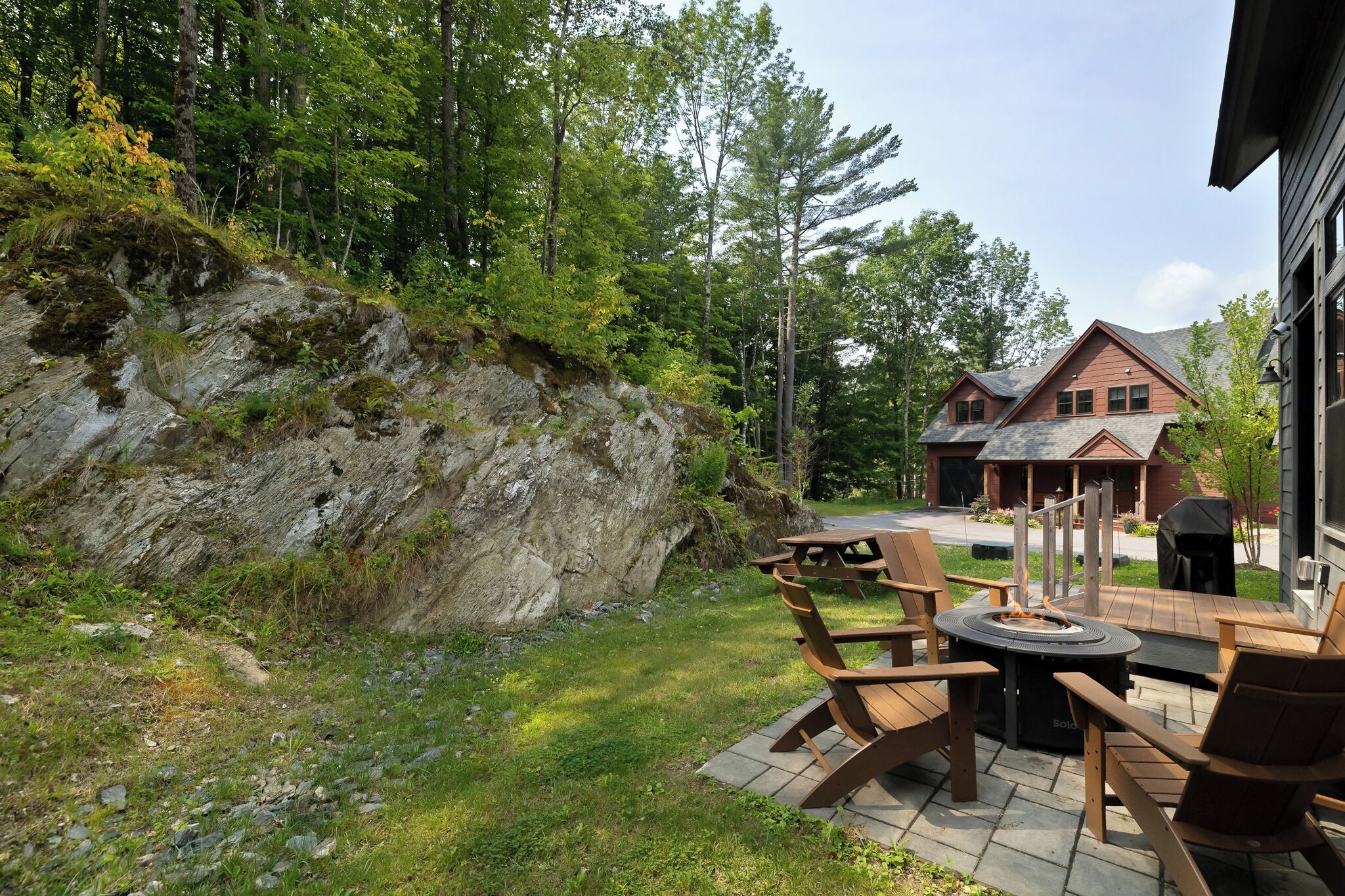 A patio with outdoor furniture and a fire pit, surrounded by a natural landscape and a house.