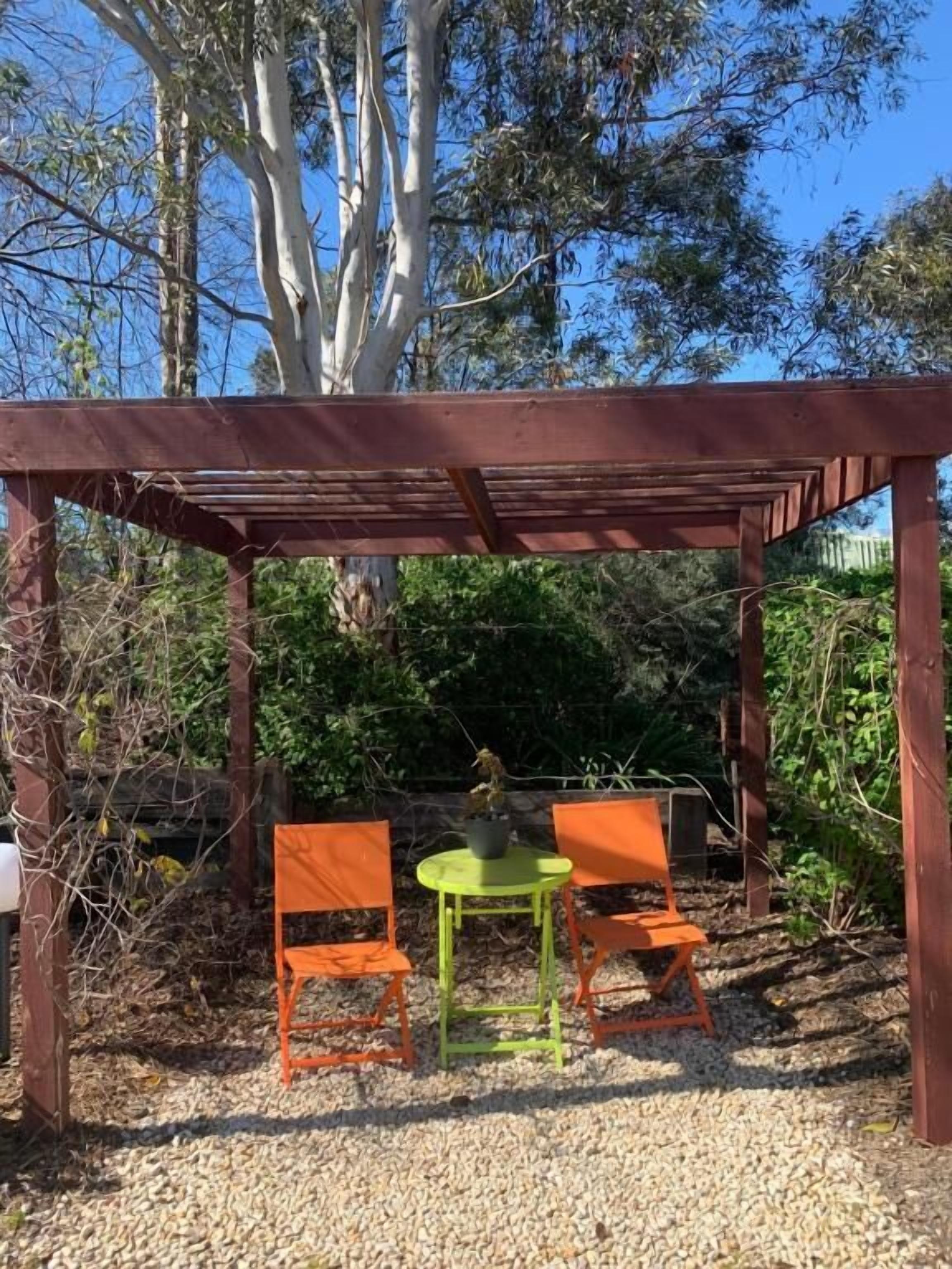 A wooden pergola with two orange chairs and a green table, surrounded by gravel and greenery.