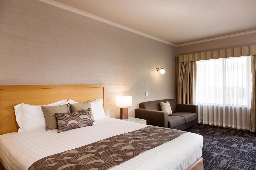 A hotel room with a large bed, a nightstand, a lamp, a sofa, and a window with curtains.