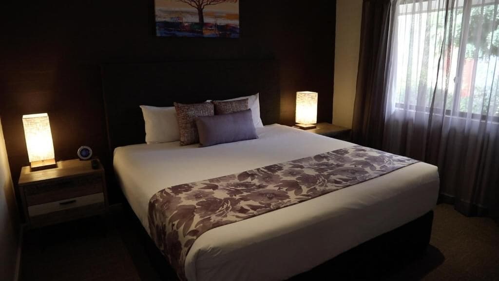 A hotel room with a large bed, two bedside tables with lamps, a painting on the wall, and a window with curtains.