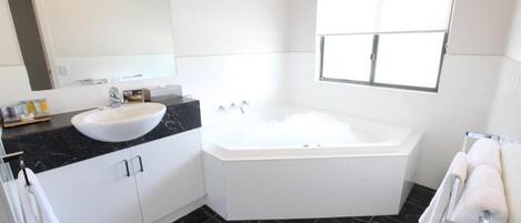 Un baño moderno con lavamanos blanco, encimera de mármol negro, un espejo grande, bañera y ventana que deja entrar luz natural.