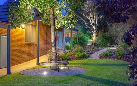 A well-maintained garden with a circular seating area, a tree, and a brick building with a window.