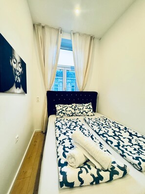 Een slaapkamer met een bed, gordijnen, een schilderij en een raam met uitzicht op gebouwen.