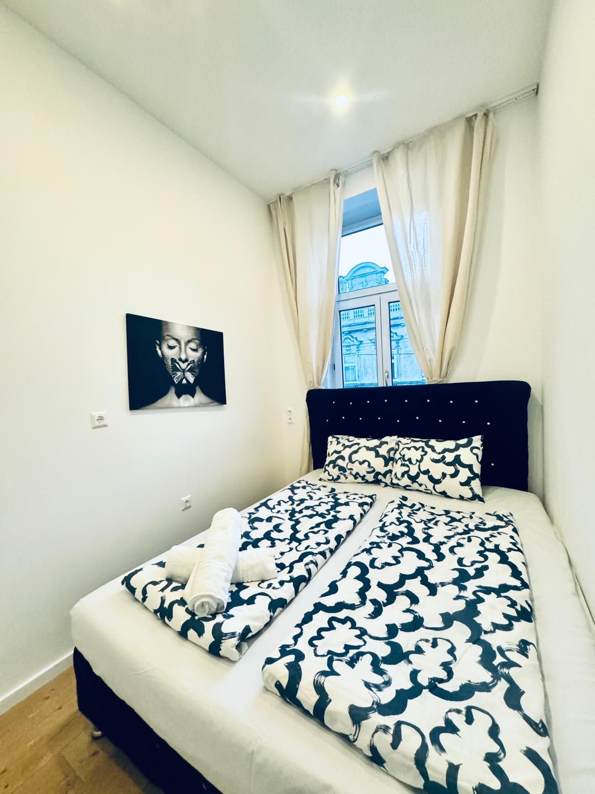 Un dormitorio con cama, ventana con cortinas, un cuadro en la pared y vista a un edificio.