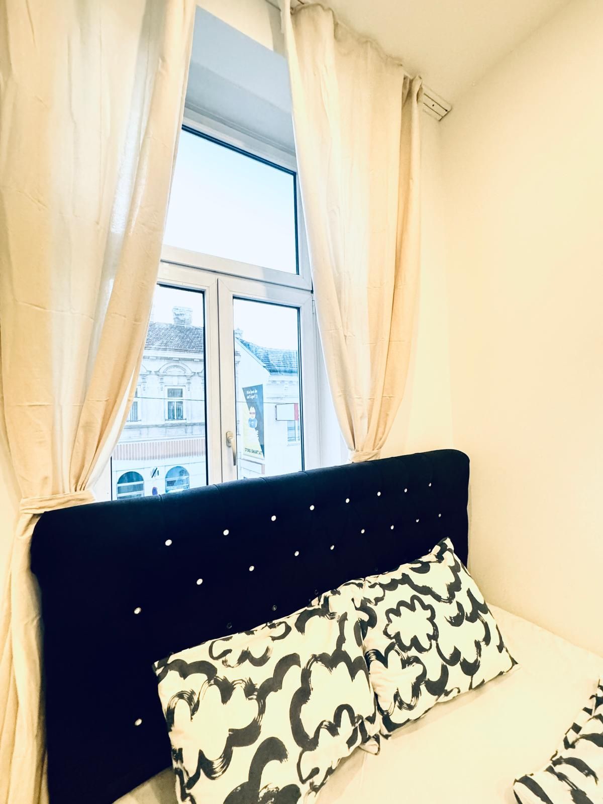 Un dormitorio con una cama, una ventana con cortinas y vistas a edificios.