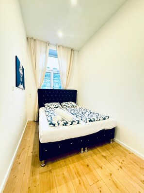 Een slaapkamer met een bed, houten vloeren, een raam met gordijnen en uitzicht op een stadsbeeld.
