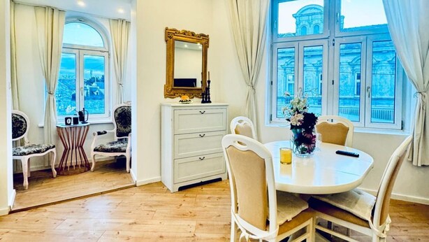 Een eethoek met een ronde tafel, stoelen, een witte dressoir, een spiegel en een raam met gordijnen.