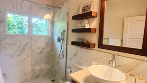 Un baño con suelo de mármol, un lavamanos blanco, un inodoro y una ducha. Hay dos ventanas con vistas a zonas verdes.