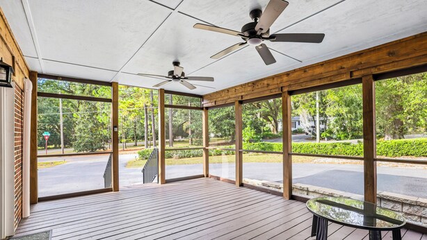 A covered outdoor area with wooden flooring, a glass table, ceiling fans, and large windows offering a view of a road and greenery.