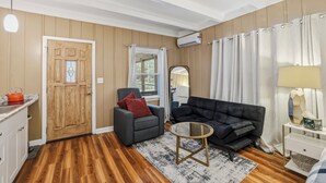 A living room with a wooden door, a grey armchair, a black sofa, a round coffee table, and a white side table with a lamp.