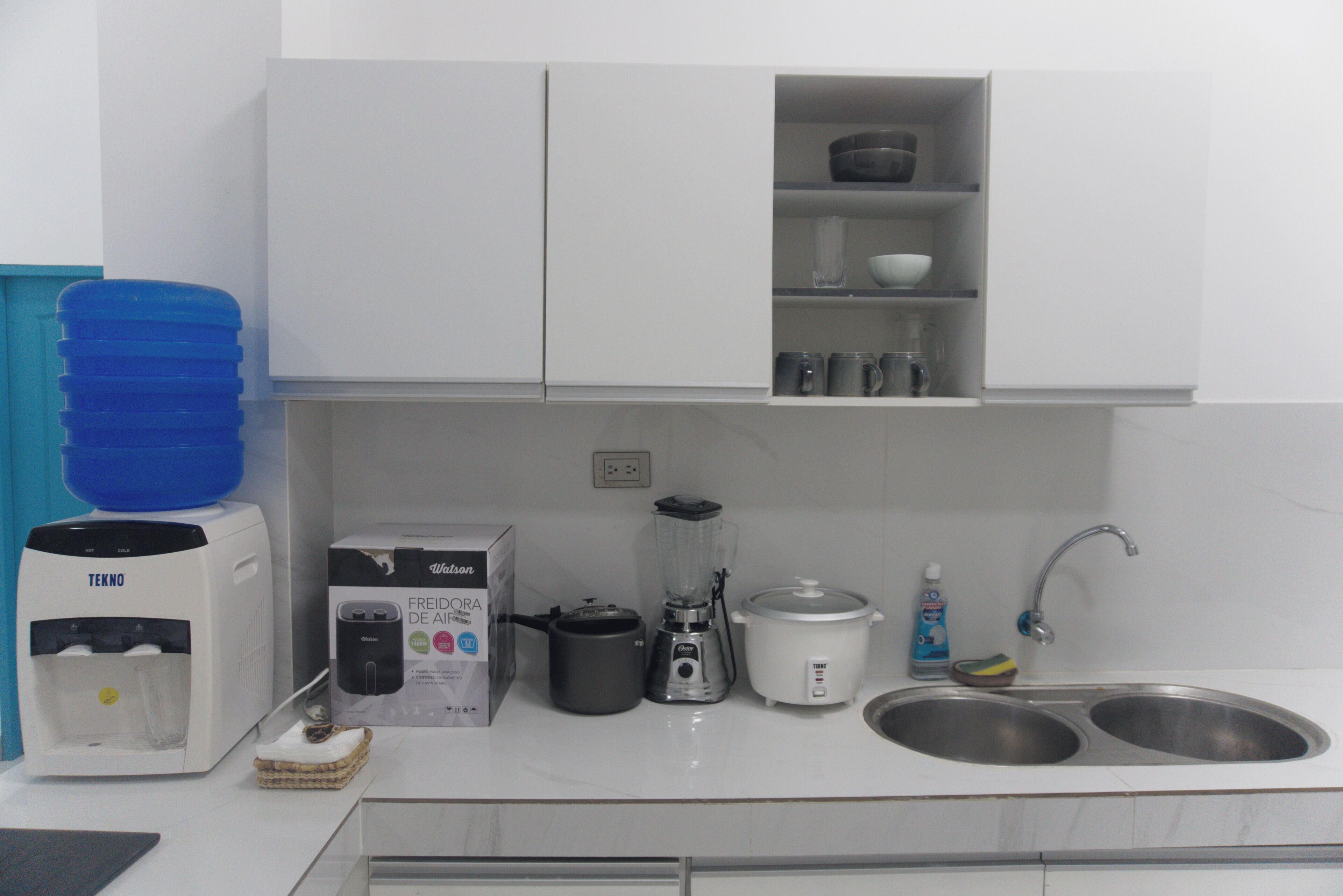 A kitchen with white cabinets, a water dispenser, a coffee maker, a blender, a slow cooker, and a sink.