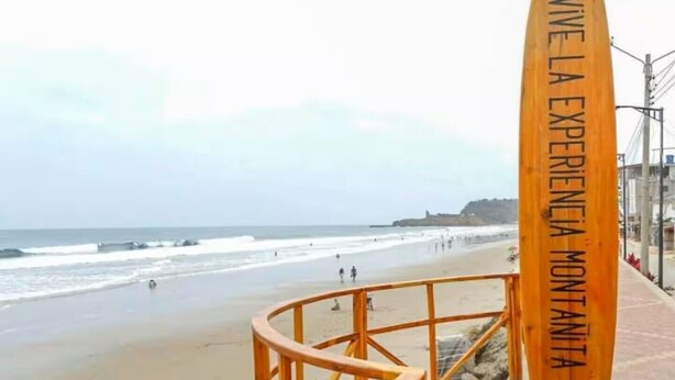 A wooden board with 'VIVE LA EXPERIENCIA MONTANITA' text, a beach with people, and a view of the ocean.