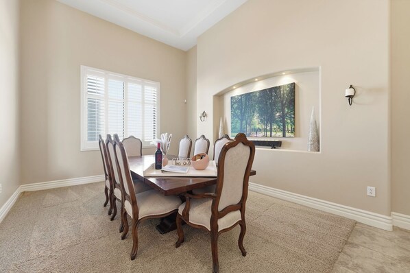 A dining room with a wooden table and chairs, a window with blinds, a painting above the window, and a wall-mounted light fixture.
