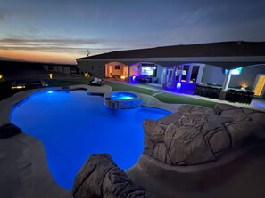 A swimming pool with a hot tub and rock features, surrounded by a patio with outdoor seating and a building in the background.