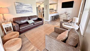 A living room with a brown leather sofa, a round wicker chair, and a small white table with a lamp. There is a kitchen area in the background with white cabinets and a black countertop. - Things are different at the beach ~~~ 🏖️ Come enjoy the breeze! (Gulf Shores)