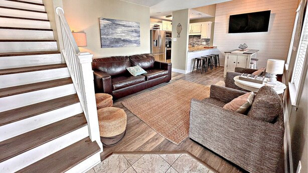 A spacious living area with a staircase, a brown leather sofa, a patterned armchair, and a kitchen in the background. - Things are different at the beach ~~~ 🏖️ Come enjoy the breeze! (Gulf Shores)