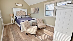 A bedroom with a large bed, two armchairs, a nightstand, a dresser, a ceiling fan, a window with blinds, and a painting on the wall. - Things are different at the beach ~~~ 🏖️ Come enjoy the breeze! (Gulf Shores)