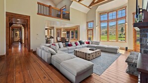 A spacious living room with a large sectional sofa, a stone fireplace, and large windows offering a view of the outdoors. - Rustic 8B Estate on 40 acres – 9,900sqft Estate on Private Ranch with 2 Hot Tubs (Heber City)