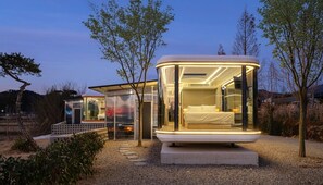 A modern, glass-walled structure with a bed and a small seating area, surrounded by trees and gravel. - Gyeongju Orion Caravan (Gyeongju)