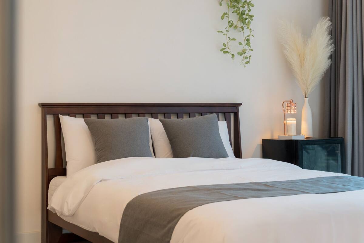 Una cama bien hecha con cabecera de madera, una mesita de noche con lámpara y una planta colgada en la pared.