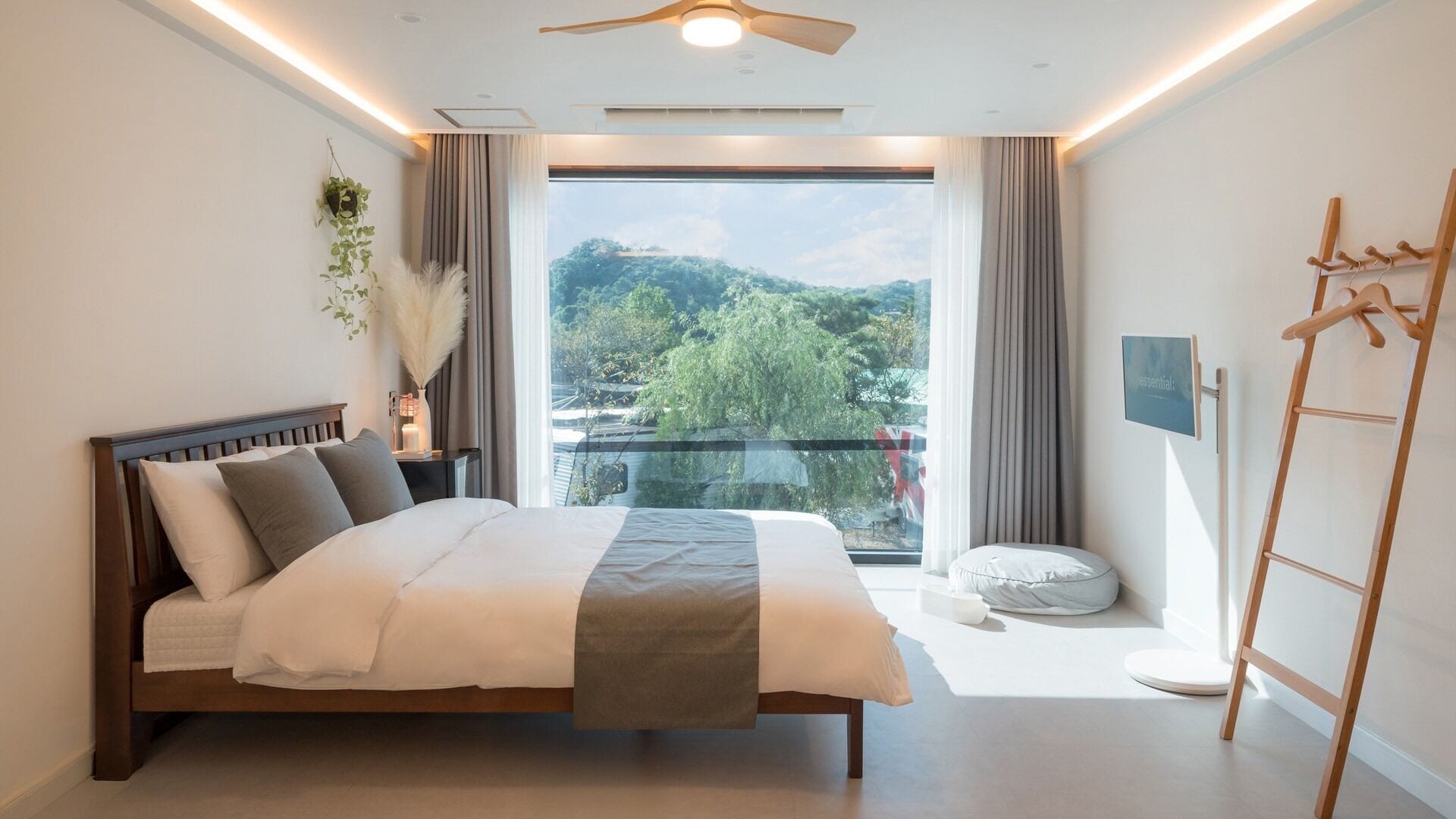 A bedroom with a bed, a ceiling fan, a window with a view of greenery, a bedside table with a lamp, and a wooden ladder.