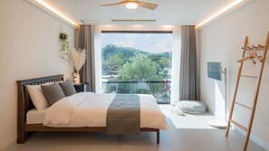 Une chambre avec un lit, un ventilateur de plafond, une fenêtre donnant sur un paysage verdoyant, une table de chevet avec une lampe et une échelle en bois.
