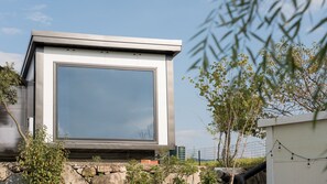 Een modern gebouw met een groot raam, omgeven door groen en een stenen muur.
