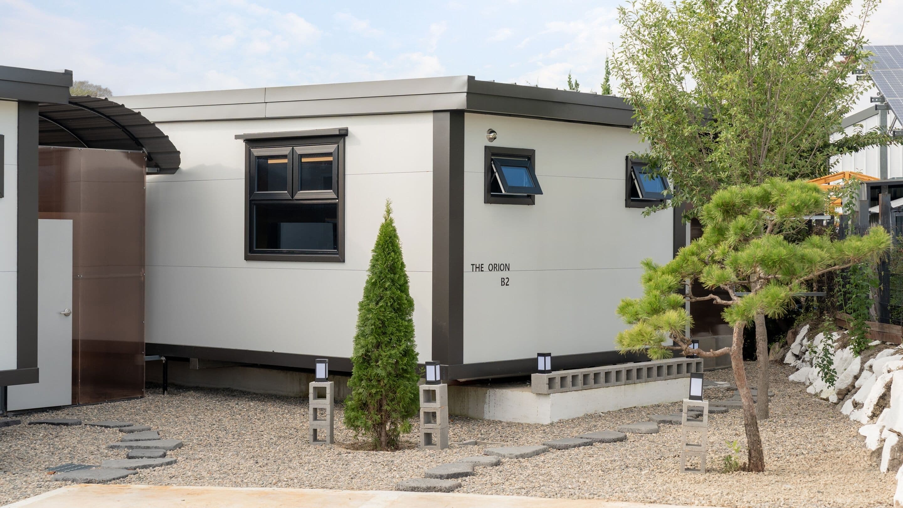 A modern building labeled "THE ORION B2" with a minimalist design, featuring a small window, a door, and a concrete pathway leading to a small tree.