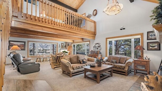 A spacious living room with a high ceiling, wooden balcony, and large windows offering views of the outdoors. - Majestic views! 4 bedroom, 4.5 bath home close to town (Estes Park)
