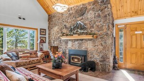 A cozy living room with a stone fireplace, wooden ceiling, and a large window overlooking trees. - Majestic views! 4 bedroom, 4.5 bath home close to town (Estes Park)