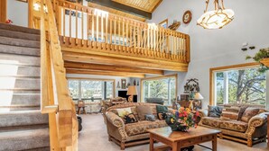 A spacious living room with a high wooden staircase, large windows, and a cozy fireplace. - Majestic views! 4 bedroom, 4.5 bath home close to town (Estes Park)