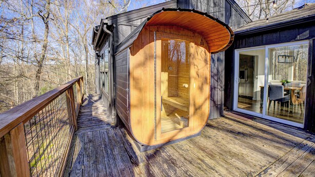 Una cabaña de madera con puerta de cristal que da a un interior de madera, un porche y vistas de una zona boscosa.