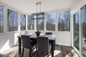 Enjoy meals in this spacious dining area with a large table and four chairs, surrounded by a high ceiling and large windows offering views of the wooded landscape.