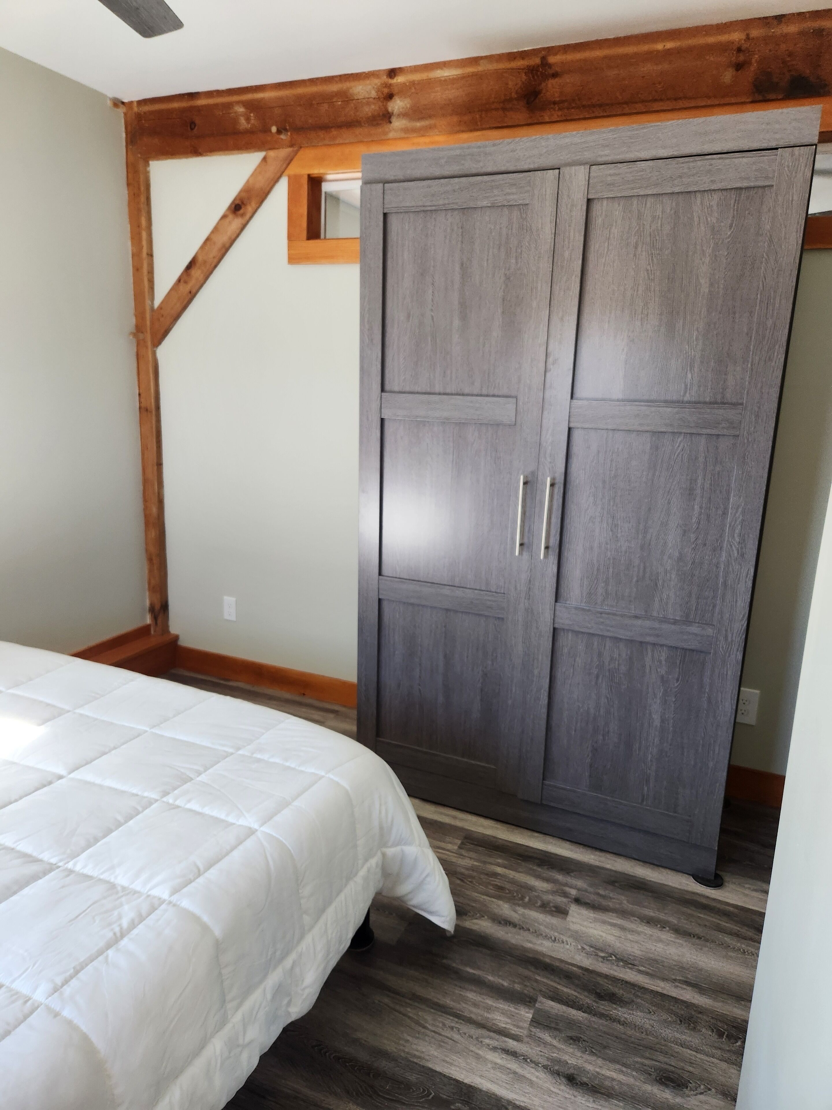 A bedroom with a bed, wooden beams, a grey wardrobe, and a white quilted bedspread.