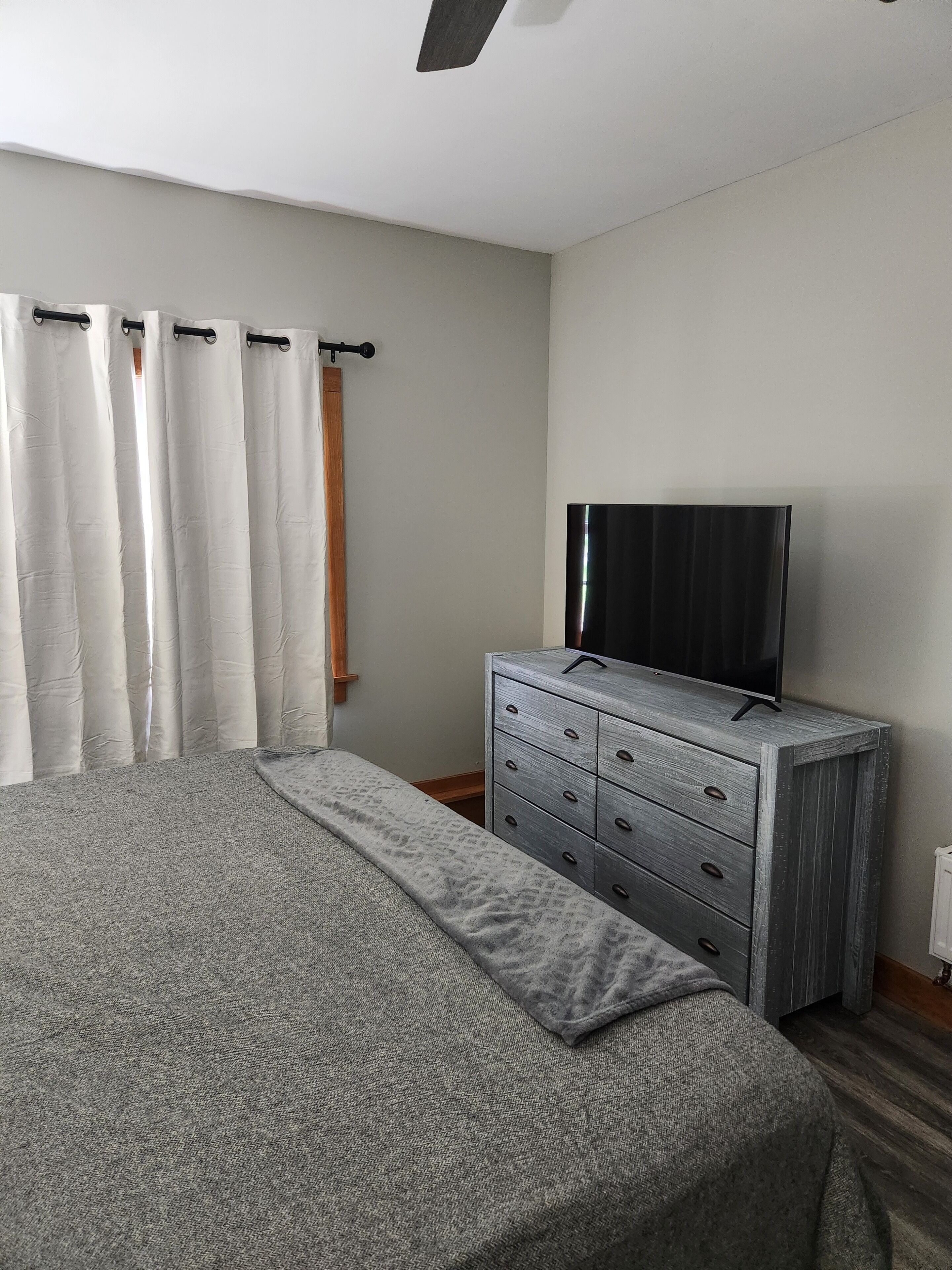 A bedroom with a bed, a dresser, a television, and a window with curtains.