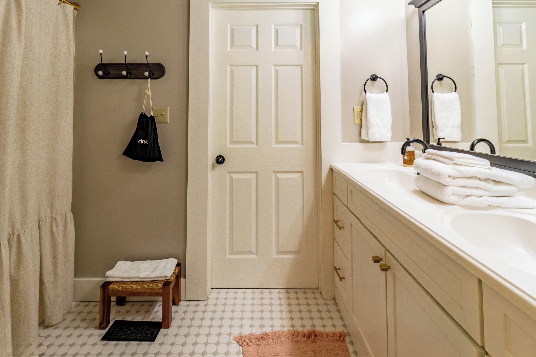 A bathroom with a white door, a towel rack with a black bag, a small wooden stool, and a white sink with a mirror.