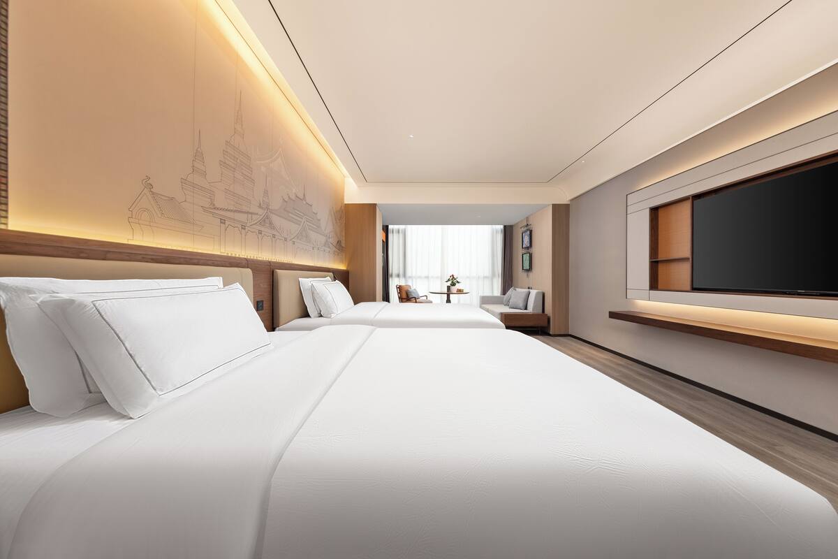 Quarto de hotel com duas camas, uma televisão de tela grande, uma escrivaninha com cadeira e uma prateleira na parede.