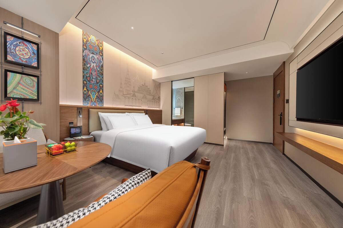 Quarto de hotel moderno com uma cama grande, uma mesa com planta, uma TV e uma parede com padrões decorativos.