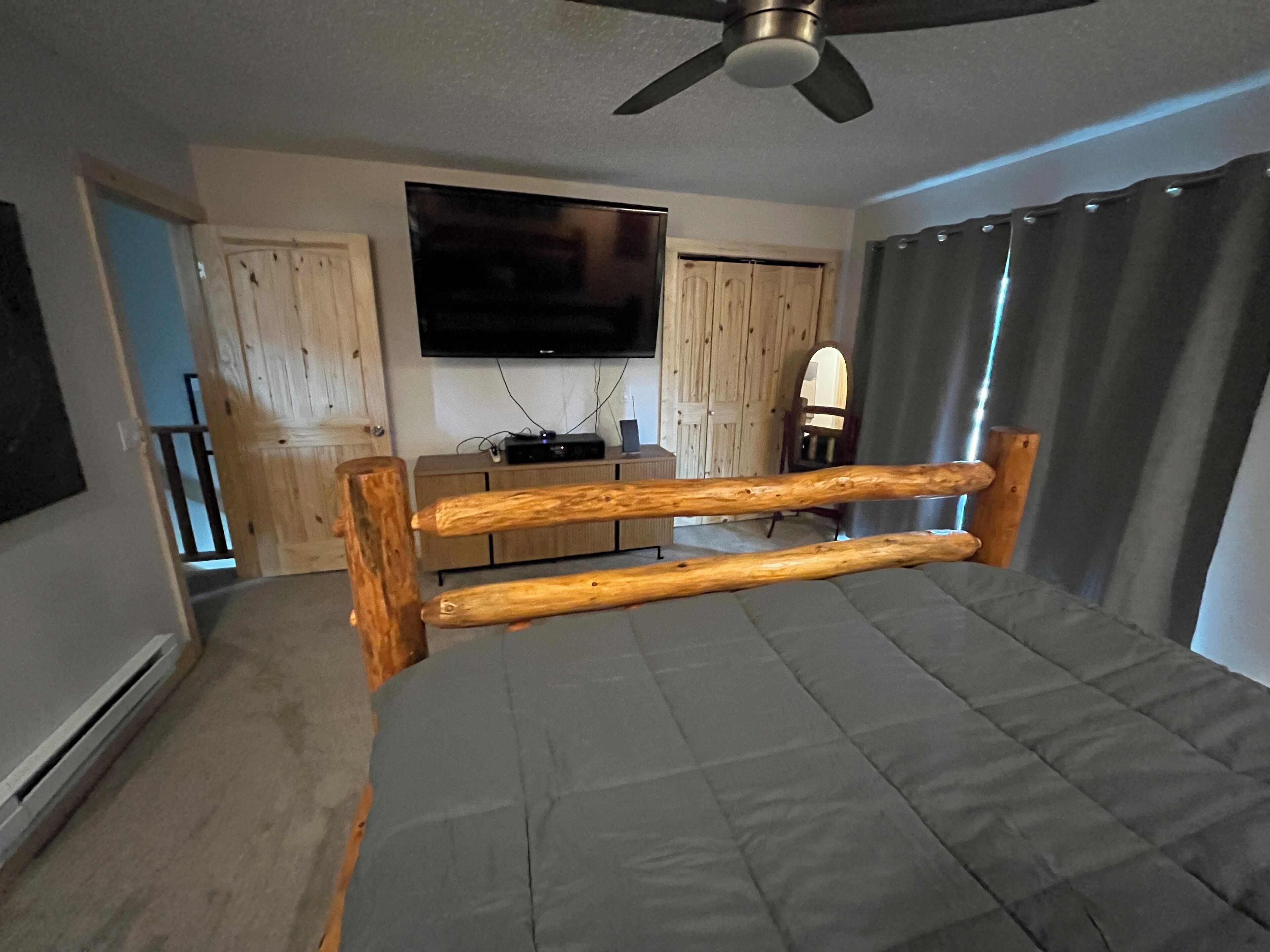 Ein Schlafzimmer mit Bett, Holz-Kopfstück, Deckenventilator, flachbildschirmfernseher an der Wand, Holzkommode und Fenster mit Vorhängen.