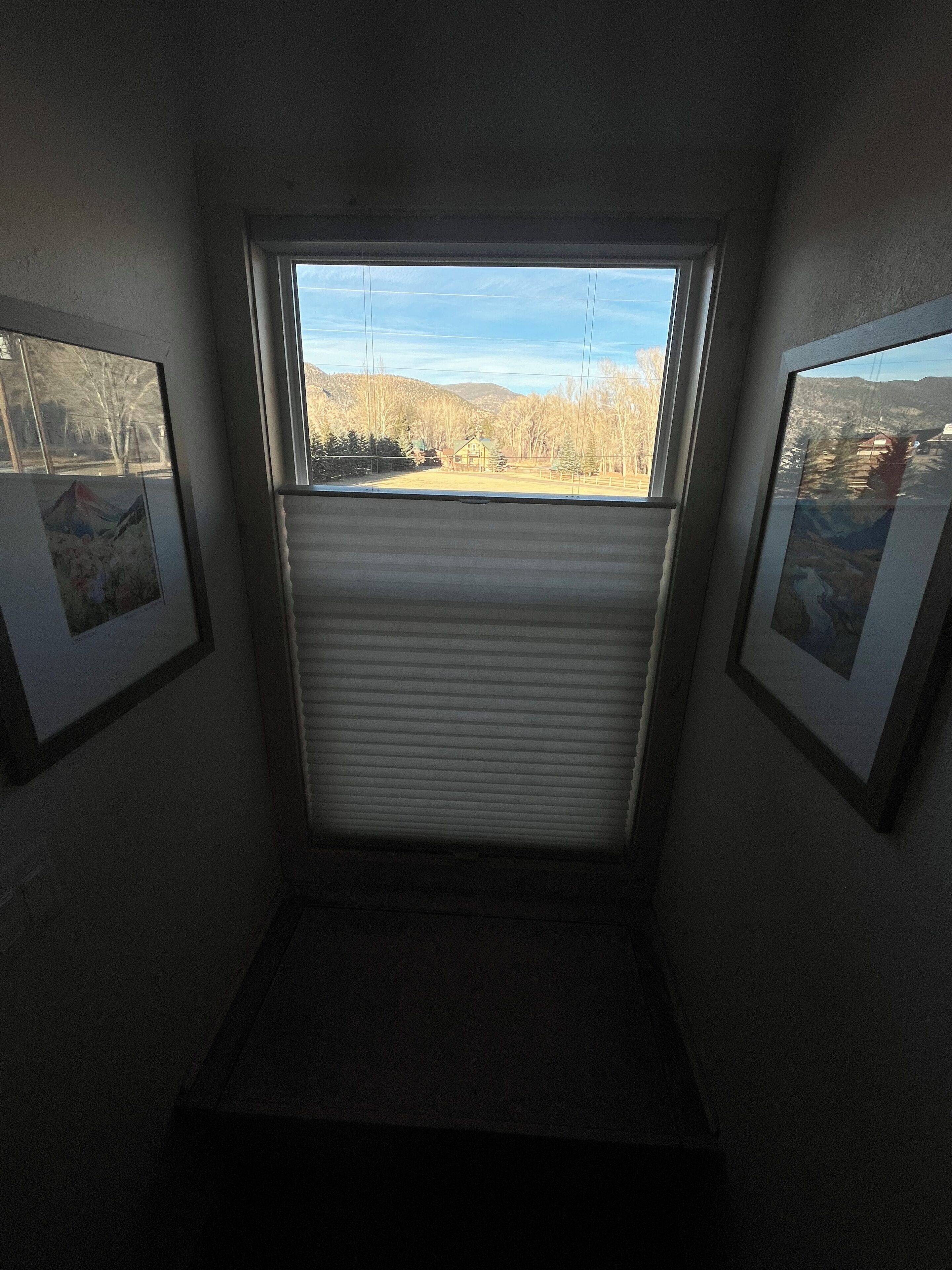 A narrow hallway with a window at the end, framed pictures on the walls, and a closed roller shutter.